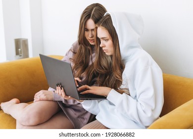 Young Women Sisters Twins Teenagers In Hoodie Beautiful Girls Using Laptop Sitting On Yellow Couch In The Modern Interior