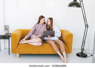 Young Women Sisters Twins Teenagers In Hoodie Beautiful Girls Using Laptop Sitting On Yellow Couch In Modern Interior