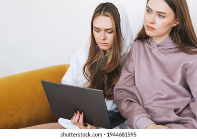 Young Women Sisters Twins Teenagers In Hoodie Beautiful Girls Using Laptop Sitting On Yellow Couch In Modern Interior