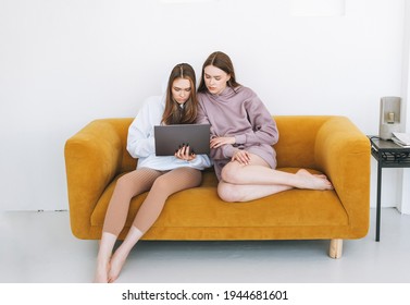 Young Women Sisters Twins Teenagers In Hoodie Beautiful Girls Using Laptop Sitting On Yellow Couch In Modern Interior