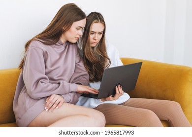 Young Women Sisters Twins Teenagers In Hoodie Beautiful Girls Using Laptop Sitting On Yellow Couch In Modern Interior