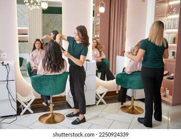 Young women sitting in chairs in front of mirrors while hairdressers styling clients hair. Two female hairstylists curling client hair and doing hairstyle in modern beauty salon. - Powered by Shutterstock