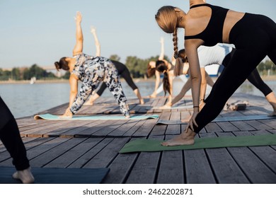 Young women stand in elongated triangle pose and do trikonasana on riverbank. Yoga class with instructor at city pier. Group of girls is engaged in physical exercises or Pilates. Outdoor training. - Powered by Shutterstock