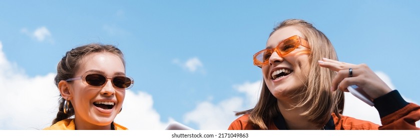 young women in trendy sunglasses smiling against blue cloudy sky, banner - Powered by Shutterstock