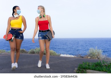 Young women walking, carrying a skateboards and looking at each other with protective mask for coronavirus. Friendship and covid concept. - Powered by Shutterstock