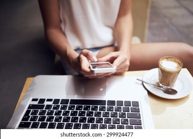 Young Women Working In Coffee Shop. Student Working With Laptop, Holding IPhone. A Cup Of Coffee On The Table 