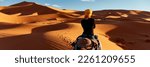 A young women in a yellow cap rides a camel  through the dunes in the Sahara Desert. View of the woman from behind, in the background, small silhouettes of other tourists. Merzouga, Morocco