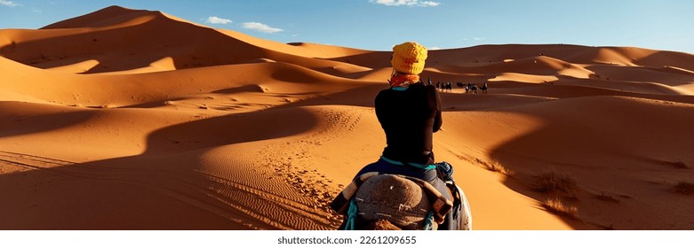 Una joven con una gorra amarilla recorre un camello por las dunas del desierto del Sahara. Vista de la mujer desde atrás, al fondo, pequeñas siluetas de otros turistas. Merzouga, Marruecos