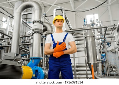 A Young Worker In A Chemical Factory Wears Orange Gloves. Metal Structures And Stainless Steel Pipes On The Background.