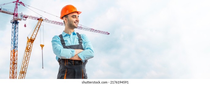 Young worker at construction site. Builder guy wearing protective workwear, hard hat, construction cranes on skyline. Construction workforce, first job working labor man.