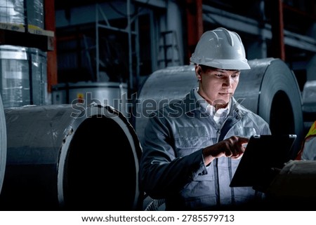 Young worker with tablet inspecting large metal tubes at factory during night shift. Engineer with helmet and safety vest working in industrial site. Inspector checking quality of product in a dark