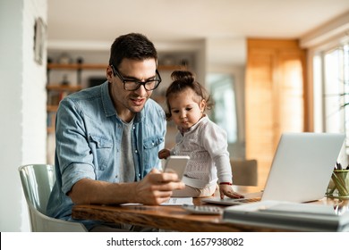 Young working father text messaging on mobile phone while being with his small daughter at home. 