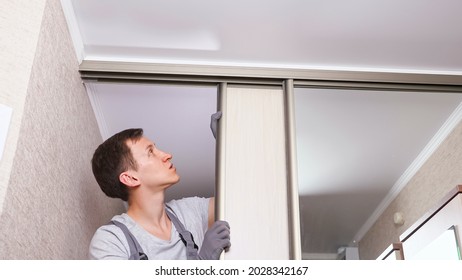 Young workman in uniform fixes wooden sliding door of contemporary built-in wardrobe with mirror in light entry room close low angle shot