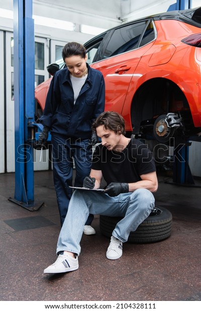 Young workman writing on\
clipboard on tire near colleague with screwdriver in car\
service