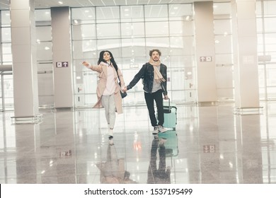 Young Worried Couple Running Together Through Empty Airport. Floor With Reflection And White Room. Guy Has Hand Of Suitcase. Woman Point Worward. Business Trip Or Vacation. Late On Flight