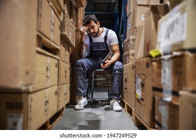 Young Worried Hardworking Tattooed Bearded Blue Collar Worker In Overalls Sitting On The Chair In Storage Of Import And Export Firm, Holding Tablet And Touching His Head. Is Delivery On Time?