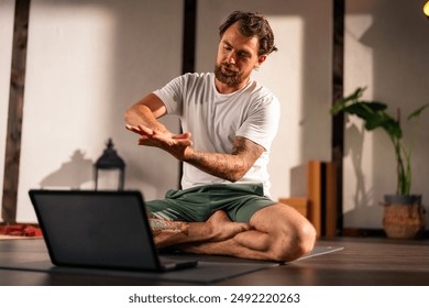 A young yoga instructor is conducting an online class while sitting on a yoga mat in his studio. He explains to his student using hand gestures. - Powered by Shutterstock