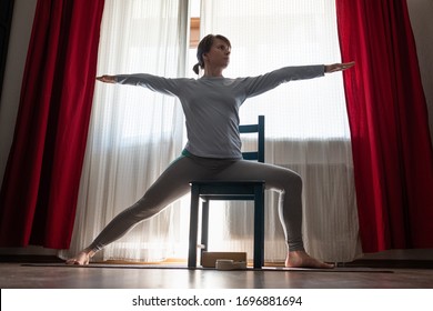 Young yoga woman doing warrior pose using chair at the living room, home. Virabhadrasana two pose.