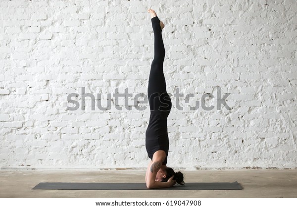 Young yogi attractive woman practicing yoga concept,
standing in salamba sirsasana exercise, headstand pose, working
out, wearing sportswear, black tank top and pants, full length,
loft background 
