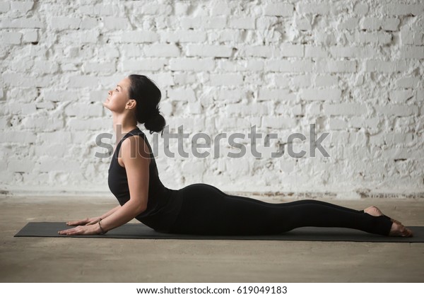 Young yogi attractive woman practicing yoga concept,\
stretching in Cobra exercise, Bhujangasana pose, working out,\
wearing sportswear, black tank top and pants, full length, white\
loft background 