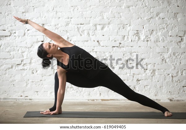 Young yogi attractive woman practicing yoga concept,\
standing in Utthita parsvakonasana exercise, Extended Side Angle\
pose, working out, wearing black sportswear, full length, white\
loft background 