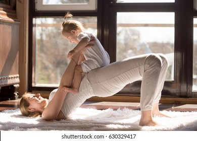 Young Yogi Smiling Mother Doing Half Bridge Exercise, Wearing White Sportswear, Working Out With Baby Daughter, Family Yoga At Home, Having No Time For Sport Club. Healthy Lifestyle Concept Photo 