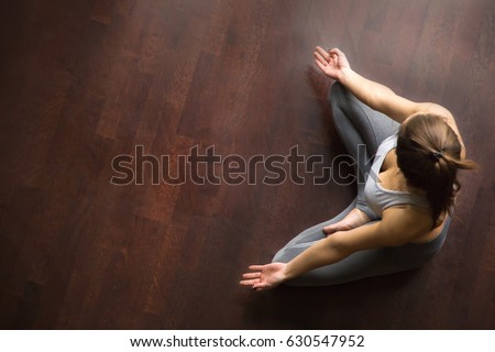 Young yogi woman practicing yoga, sitting in Half Lotus exercise, Ardha Padmasana pose, working out, wearing sportswear, grey pants, indoor, home interior, wooden floor, high angle view, copy space 