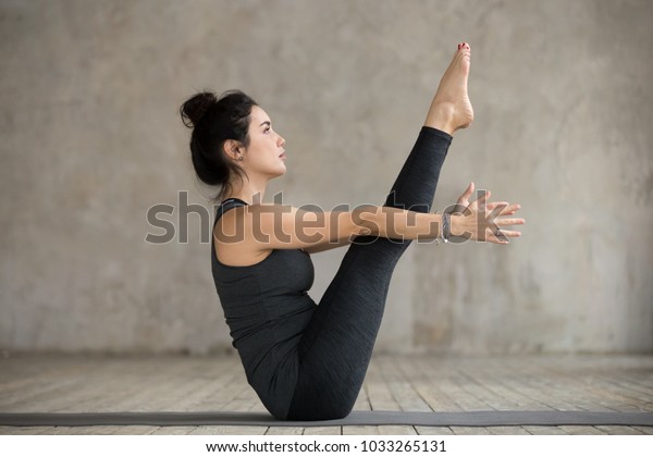 Young yogi woman\
practicing yoga, doing Paripurna Navasana exercise, boat pose,\
working out, wearing sportswear, black pants and top, indoor full\
length, gray wall in yoga\
studio