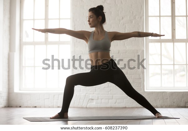 Young yogi woman practicing yoga, standing in\
Warrior two exercise, Virabhadrasana II pose, working out wearing\
sportswear bra and pants, full length, white loft studio\
background. Weight loss\
concept