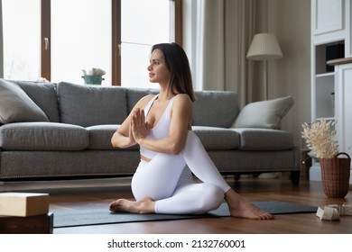 Young yogini woman practising asanas at home, wear white sportswear stretch body performs Half Lord of the Fishes pose Ardha Matsyendrasana exercise with namaste sign. Yoga, healthy lifestyle concept