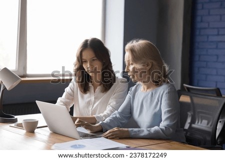 Younger 35s and older 60s women colleagues having common task work together sit at desk use laptop, talk, discuss on-line project, learn software or corporate program. Teamwork, tech, apprenticeship