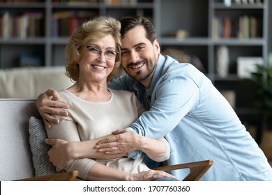 Younger And Older Generation Relatives People Portrait, Love, Care, Connection, Family Bonds Concept. Grown Up Adult Son Hugs Elderly Mother While She Resting Seated On Cozy Comfort Armchair Indoors