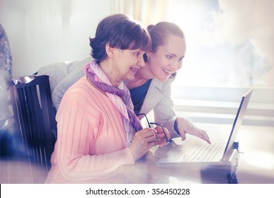 Younger Woman Helping An Elderly Person Using Laptop Computer For Internet Search. Young And Pension Age Generations Working Together.