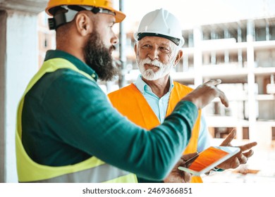 A younger worker in a green vest and hard hat shows a tablet to an older site manager in white, both engaged in active dialogue at a bustling construction location. - Powered by Shutterstock