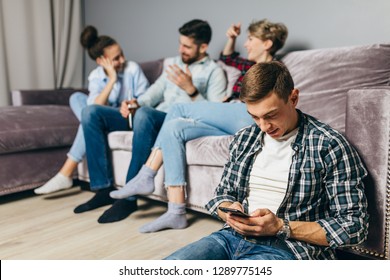 Youny Pleasant Man In Checked T-shirt Is Using His Mobile Phone While His Friends Are Having Fun. Odd Man In The Company