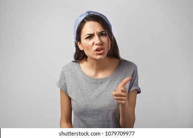 It's your fault. Studio shot of disappointed angry young brunette woman frowning wearing headscarf and gray t-shirt, pointing index finger at camera, being mad, blaming all her failures on you