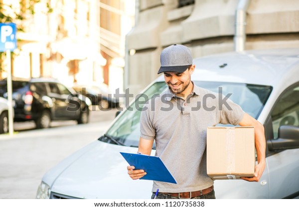 your shipping is here.\
delivery man in grey shirt with cap standing with his cardboard box\
looking on the form for last details, in front of his car, outside\
on the street