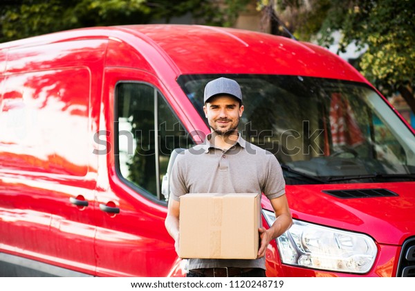 your shipping is\
here. happy delivery man in grey shirt with cap standing with his\
cardboard box on the street looking to camera with smile, in front\
of his delivery car