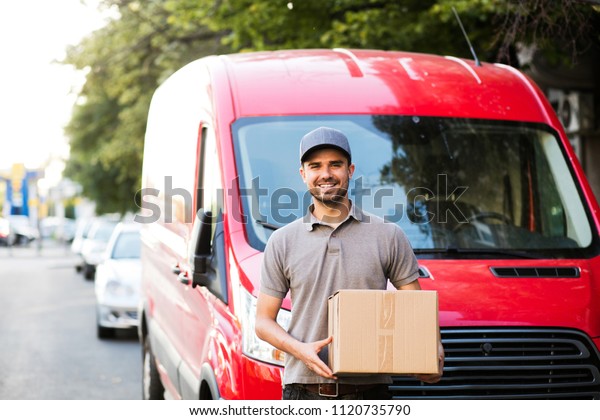 your shipping is\
here. happy delivery man in grey shirt with cap standing with his\
cardboard box on the street looking to camera with smile, in front\
of his delivery car
