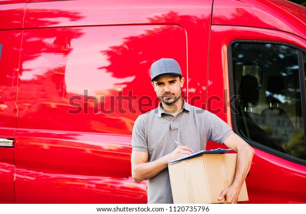 your shipping is here. young delivery man in grey\
shirt with cap standing with his cardboard box completing the form\
for deliver, ready to meet the client, outside on the street in\
front of his car