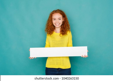 Your text here. Young excited redhead woman holding blank white banner with copy space, standing on blue studio background