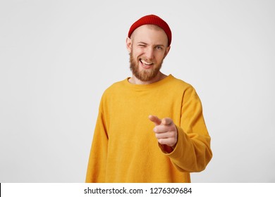 You're chosen. A young attractive interesting unusual guy dressed in a yellow sweater and a red hat winks with one eye, his finger points to the camera, encouraging, an approving gesture.