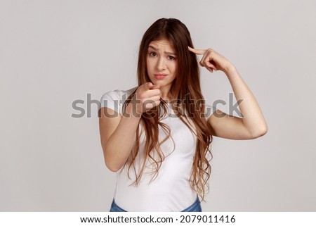 You're idiot with insane crazy plan. Displeased woman showing stupid gesture and pointing to camera, accusing of dumb idea, wearing white T-shirt. Indoor studio shot isolated on gray background.