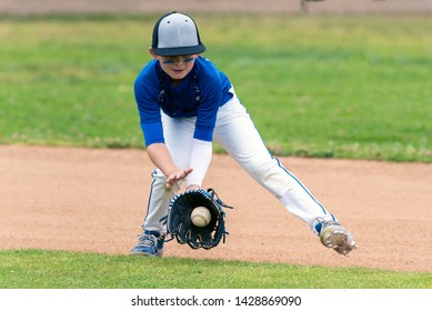Youth baseball player in blue uniform fielding a ground ball into his glove in the infield during a game.