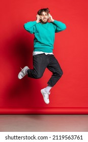 Youth Culture. Portrait Of Young Calm Man Listening To Music In Headphones And Jumping Isolated Over Red Studio Background. Concept Of Youth, Emotion, Facial Expression, Fashion, Lifestyle, Ad