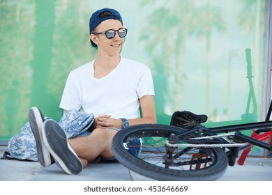 Youth And Happiness. Happy 15-year Old Student Wearing Snapback And Shades, Smiling, Looking Away, Enjoying Summer Vacations, Sitting In The Shadow With His Fixed Gear Bicycle Lying Beside Him