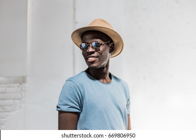 Youth Lifestyle And Fashion. Handsome Afro American Man Model Wearing Fashionable Round Shades And Beige Hat With Blue T-shirt Posing And Smiling In White Cafe With Sunlight.