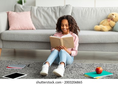 Youth and literature. Smiling african american gen z girl holding and reading paper book. Schoolgirl sitting on the floor carpet at home in cozy living room, leaning on couch