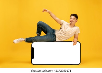 Youth Modern Lifestyle. Stylish Young Man Lying On Huge 3d Model Of Cellphone With Blank White Screen Isolated On Yellow Background, New App Or Website Ad, Copy Space For Your Ads, Mockup Or Design