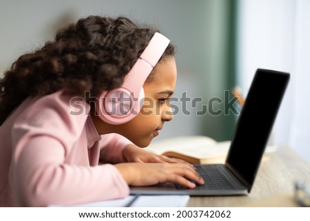 Youth and technology concept. Focused black schoolgirl using laptop computer, sitting at table too close to pc, side view. Little girl with poor eyesight having near vision problem